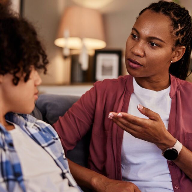 A beautiful African-American woman talking with her teenage son at home. Mother sitting on a couch having a supportive conversation with her teenage son