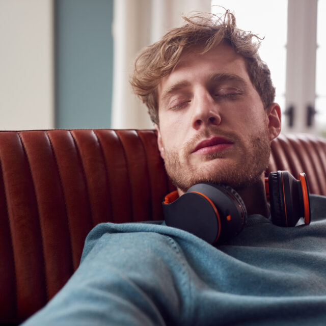 Man With Wireless Headphones Around Neck Relaxing On Armchair At Home With Closed Eyes Man resting on a couch with eyes closed and headphones around his neck