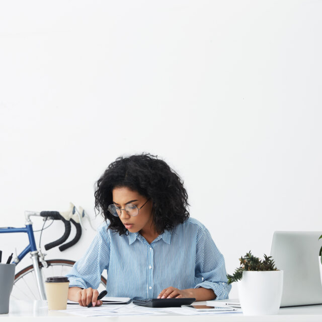 woman-working-at-desk-with-bike-and-plants-2026-09-24-20-41-58-utc Woman focused on paperwork and a calculator at her desk